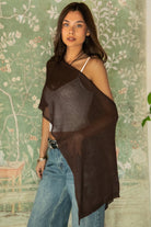 Woman wearing TIC TOC TC31873 SOLEI lightweight open sheer sweater knit poncho in BROWN, featuring a wide off-shoulder neckline, airy open knit texture, relaxed draped poncho silhouette, and asymmetrical hem, styled over a white cami with light wash denim for a western-casual summer layering look.