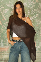 Woman wearing TIC TOC TC31873 SOLEI lightweight open sheer sweater knit poncho in BROWN, featuring a wide off-shoulder neckline, airy open knit texture, relaxed draped poncho silhouette, and asymmetrical hem, styled over a white cami with light wash denim for a western-casual summer layering look.