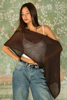 Woman wearing TIC TOC TC31873 SOLEI lightweight open sheer sweater knit poncho in BROWN, featuring a wide off-shoulder neckline, airy open knit texture, relaxed draped poncho silhouette, and asymmetrical hem, styled over a white cami with light wash denim for a western-casual summer layering look.