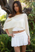 Woman wearing TIC TOC TC31873 SOLEI lightweight open sheer sweater knit poncho in CREAM, featuring a wide off-shoulder neckline, airy open knit texture, relaxed draped poncho silhouette, and asymmetrical hem, styled over a white cami with light wash denim for a western-casual summer layering look.