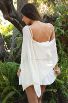 Woman wearing TIC TOC TC31873 SOLEI lightweight open sheer sweater knit poncho in CREAM, featuring a wide off-shoulder neckline, airy open knit texture, relaxed draped poncho silhouette, and asymmetrical hem, styled over a white cami with light wash denim for a western-casual summer layering look.