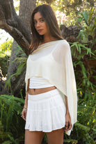 Woman wearing TIC TOC TC31873 SOLEI lightweight open sheer sweater knit poncho in CREAM, featuring a wide off-shoulder neckline, airy open knit texture, relaxed draped poncho silhouette, and asymmetrical hem, styled over a white cami with light wash denim for a western-casual summer layering look.