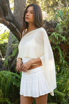 Woman wearing TIC TOC TC31873 SOLEI lightweight open sheer sweater knit poncho in CREAM, featuring a wide off-shoulder neckline, airy open knit texture, relaxed draped poncho silhouette, and asymmetrical hem, styled over a white cami with light wash denim for a western-casual summer layering look.