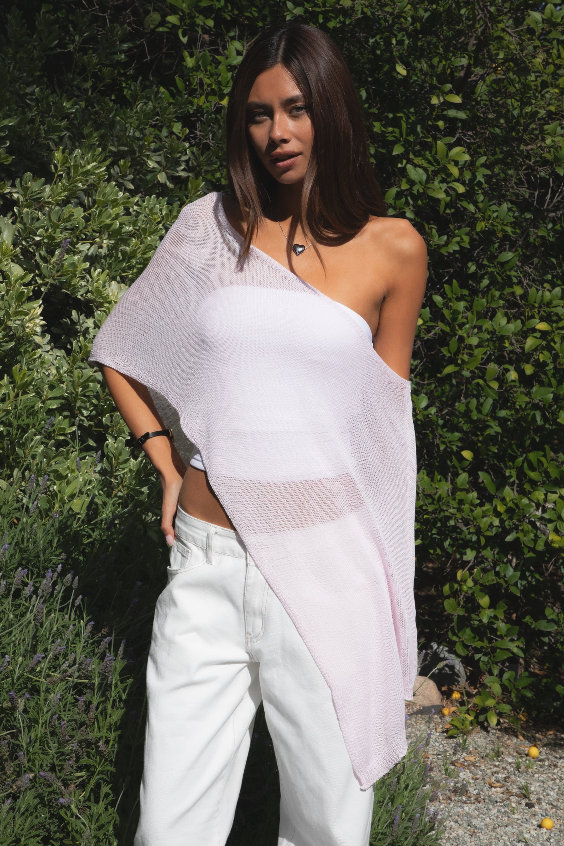 Woman wearing TIC TOC TC31873 SOLEI lightweight open sheer sweater knit poncho in PINK, featuring a wide off-shoulder neckline, airy open knit texture, relaxed draped poncho silhouette, and asymmetrical hem, styled over a white cami with light wash denim for a western-casual summer layering look.