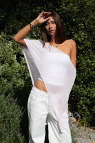 Woman wearing TIC TOC TC31873 SOLEI lightweight open sheer sweater knit poncho in PINK, featuring a wide off-shoulder neckline, airy open knit texture, relaxed draped poncho silhouette, and asymmetrical hem, styled over a white cami with light wash denim for a western-casual summer layering look.