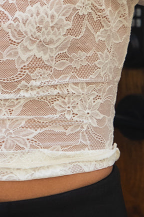 Woman wearing TIC TOC TC31877-ELAN ELAN lace off the shoulder top featuring a soft draped off-shoulder neckline, short flutter sleeves with lace trim, sheer floral lace mesh body with a fitted stretch silhouette, cropped length, and fully unlined see-through finish, styled with high-rise bottoms for a coquette-inspired going-out look.