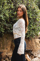 Woman wearing TC31934-VELORA floral lace off-shoulder top with bell sleeves and cropped slim fit body, styled with high-rise bottoms for a boho Y2K going-out look.