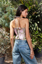 Woman wearing CAMOLETTE camo lace up halter corset top featuring stretch twill camo fabric, allover camo print, studded trim details, front lace up design with grommet accents, halter neck tie, structured corset silhouette, studded neckline and hem, and back zipper closure.