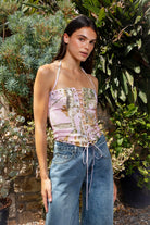 Woman wearing CAMOLETTE camo lace up halter corset top featuring stretch twill camo fabric, allover camo print, studded trim details, front lace up design with grommet accents, halter neck tie, structured corset silhouette, studded neckline and hem, and back zipper closure.