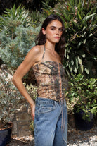 Woman wearing CAMOLETTE camo lace up halter corset top featuring stretch twill camo fabric, allover camo print, studded trim details, front lace up design with grommet accents, halter neck tie, structured corset silhouette, studded neckline and hem, and back zipper closure.