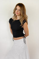 Woman wearing SELENE boat neck cowl back side tie crop top featuring an elegant boat neckline, draped cowl back, adjustable back strap, side tie ruching details, cap short sleeves, and lightweight stretch jersey fabric for a feminine cropped silhouette.