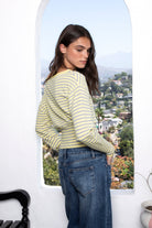 Woman wearing BELLAMY roll neck stripe off shoulder sweater top featuring soft stripe sweater knit fabric, off shoulder neckline, relaxed long sleeve silhouette, cropped hem, wide ribbed waistband, and smooth stretch feel for cozy lounge and travel styling.