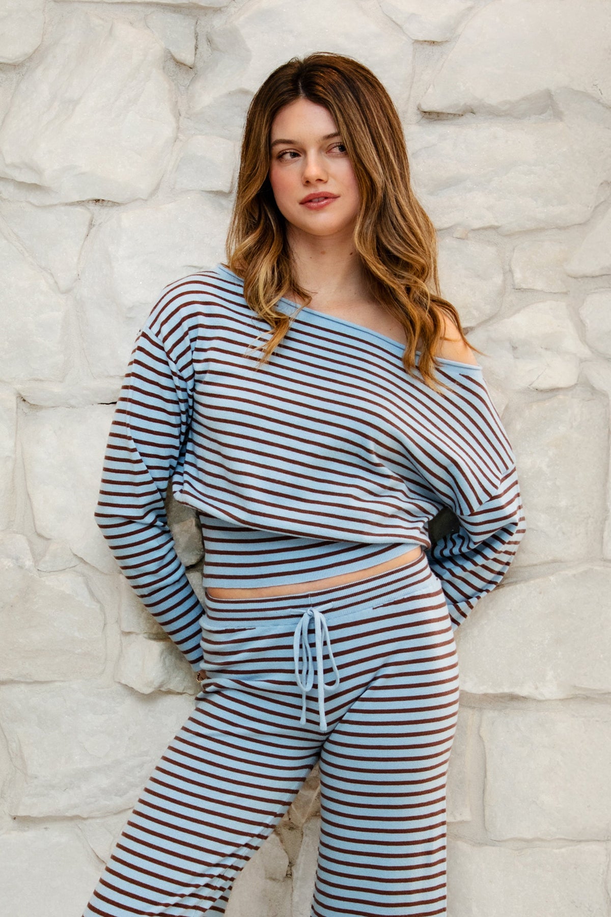 Woman wearing BELLAMY roll neck stripe off shoulder sweater top featuring soft stripe sweater knit fabric, off shoulder neckline, relaxed long sleeve silhouette, cropped hem, wide ribbed waistband, and smooth stretch feel for cozy lounge and travel styling.