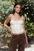 Woman wearing ELISHA ruffled lace up chiffon cropped corset top featuring softly textured chiffon fabric, gathered bust, delicate ruffle trim neckline, adjustable self tie shoulder straps, vertical ruffle detailing, fitted cropped bodice, open back with adjustable lace up closure, and soft jersey lining.