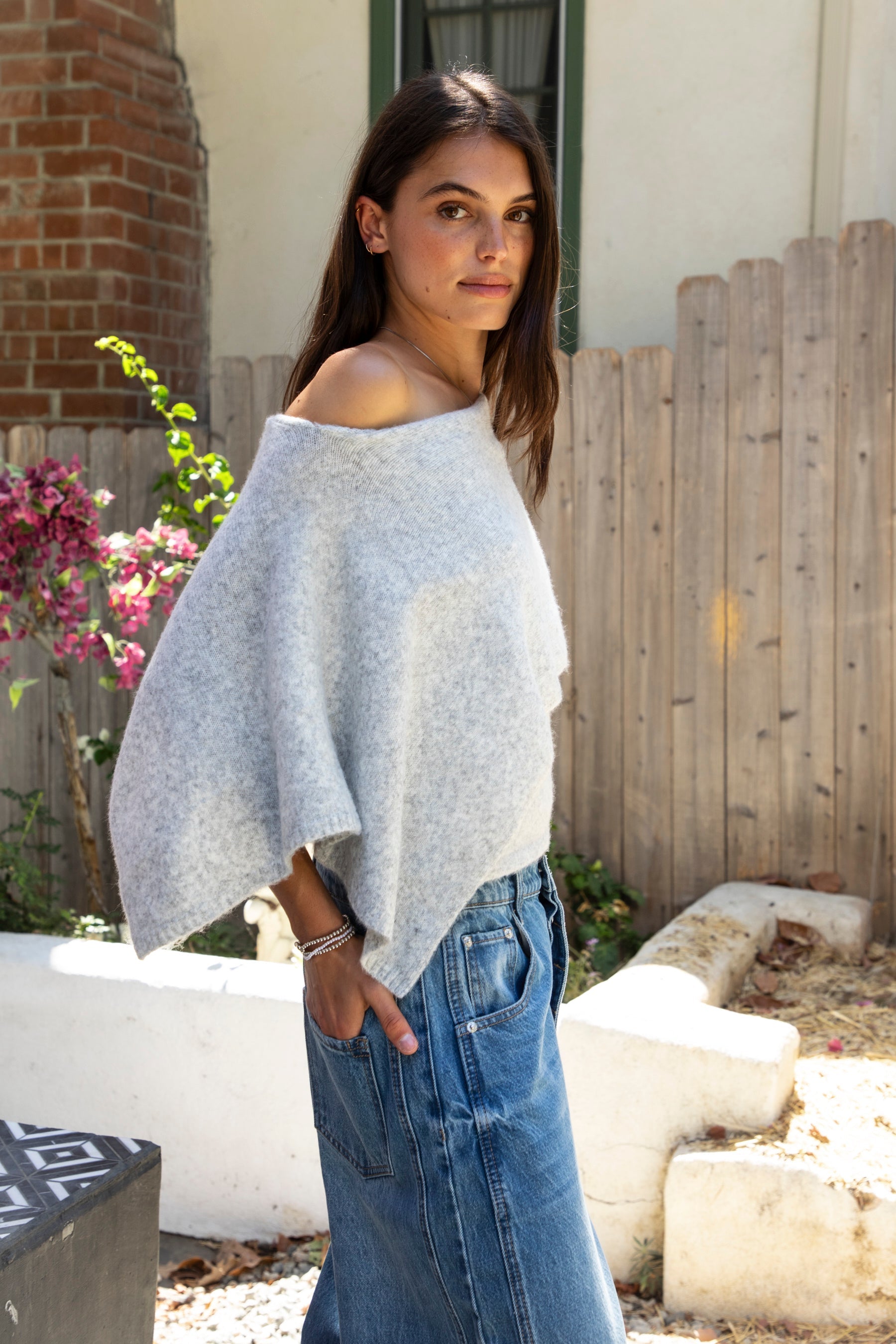 Woman wearing AINSLEY off shoulder asymmetric knit poncho sweater featuring soft brushed sweater knit fabric, cozy warmth, convertible off shoulder neckline, draped poncho silhouette, relaxed asymmetrical design, easy pull on style, and soft stretch comfort for fall and winter layering.