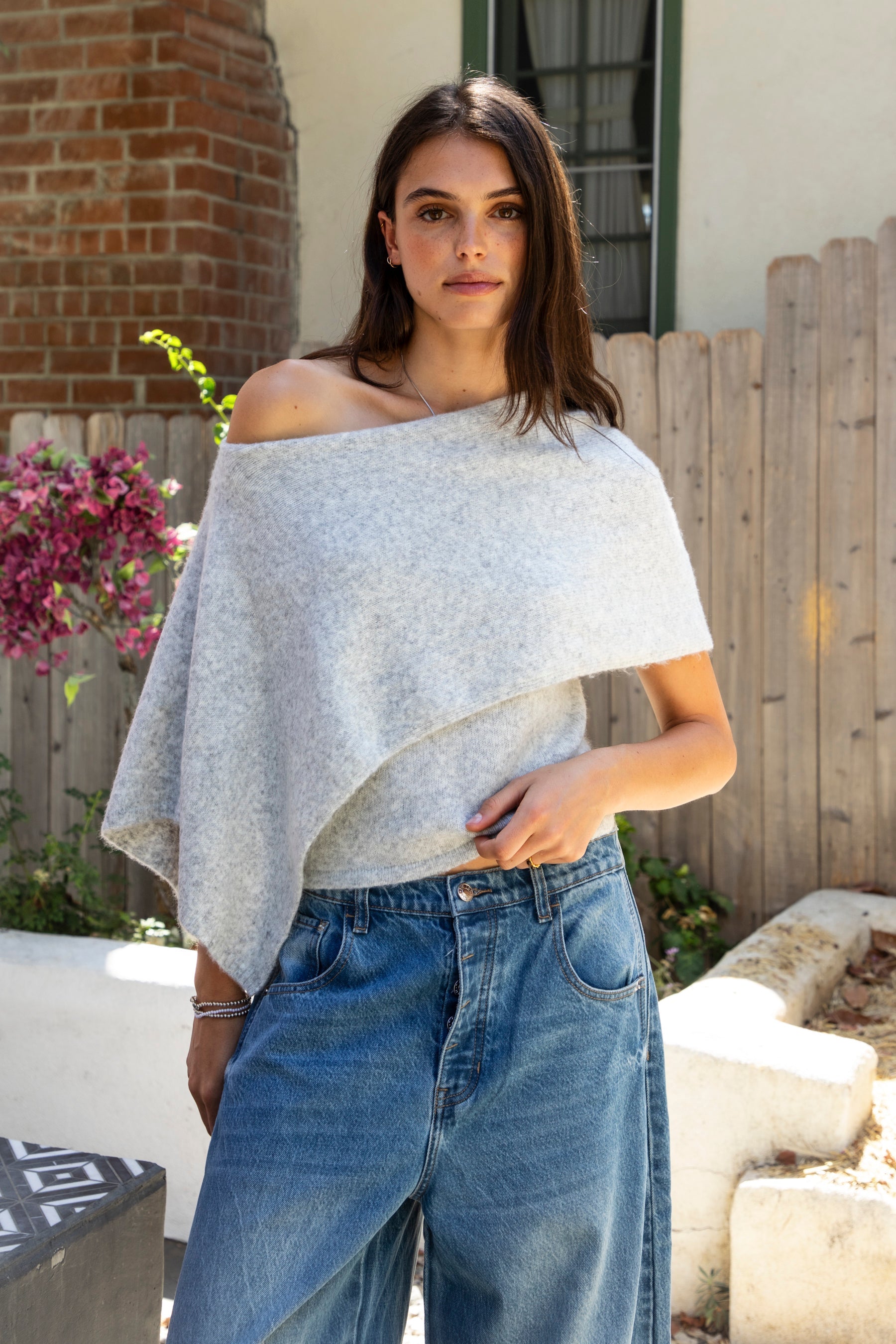 Woman wearing AINSLEY off shoulder asymmetric knit poncho sweater featuring soft brushed sweater knit fabric, cozy warmth, convertible off shoulder neckline, draped poncho silhouette, relaxed asymmetrical design, easy pull on style, and soft stretch comfort for fall and winter layering.