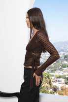 Woman wearing LORELEI sheer lace long sleeve V neck wrap top featuring sheer stretch lace, delicate textured semi sheer finish, flattering wrap front silhouette, adjustable self wrap design, fitted cropped shape, and scalloped edges along the neckline, cuffs, and hem.
