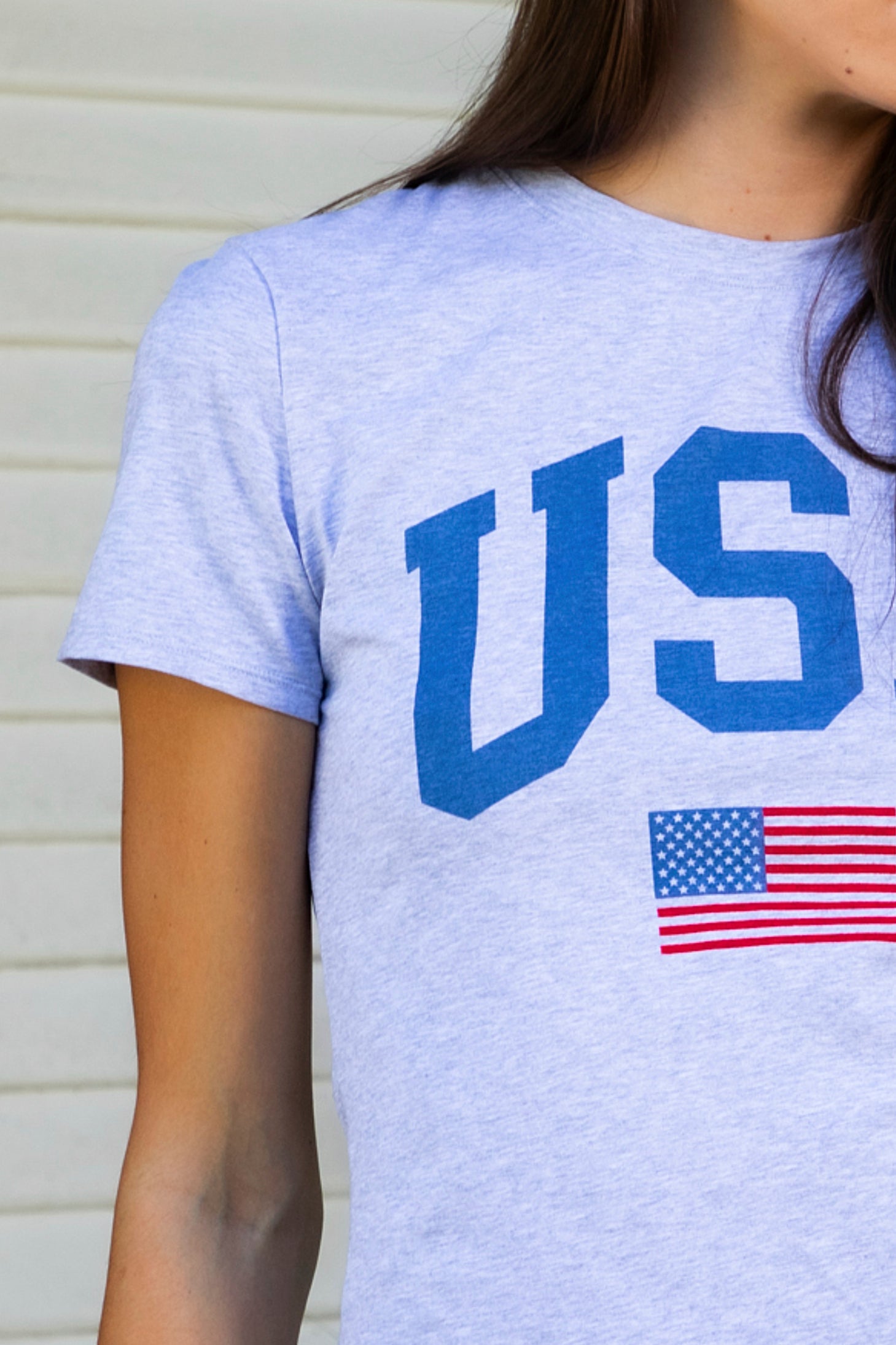 Woman wearing LIBERTY classic USA graphic crewneck baby tee featuring soft single jersey cotton stretch fabric, fitted short sleeve baby tee silhouette, clean cropped length, classic crew neckline, bold USA graphic across the front, American flag graphic detail below, and straight fitted hemline.