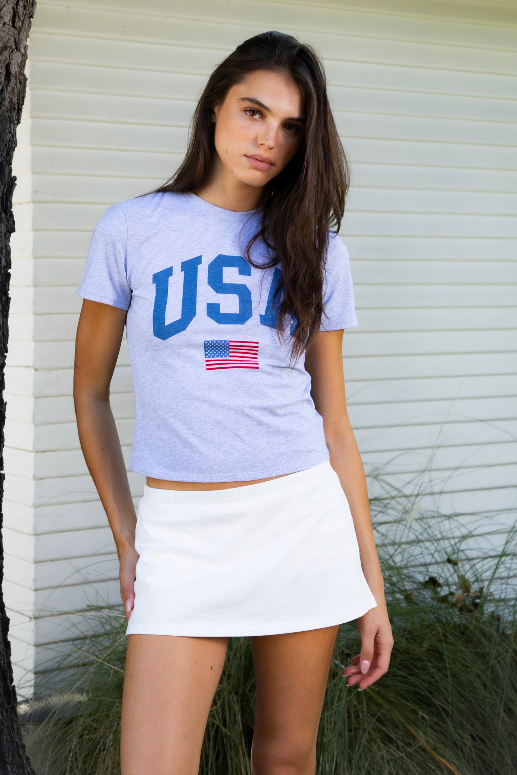 Woman wearing LIBERTY classic USA graphic crewneck baby tee featuring soft single jersey cotton stretch fabric, fitted short sleeve baby tee silhouette, clean cropped length, classic crew neckline, bold USA graphic across the front, American flag graphic detail below, and straight fitted hemline.
