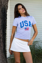 Woman wearing LIBERTY classic USA graphic crewneck baby tee featuring soft single jersey cotton stretch fabric, fitted short sleeve baby tee silhouette, clean cropped length, classic crew neckline, bold USA graphic across the front, American flag graphic detail below, and straight fitted hemline.