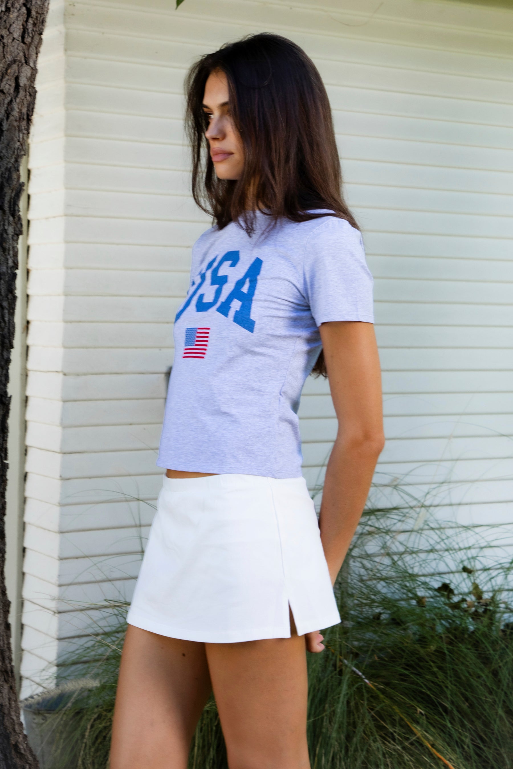 Woman wearing LIBERTY classic USA graphic crewneck baby tee featuring soft single jersey cotton stretch fabric, fitted short sleeve baby tee silhouette, clean cropped length, classic crew neckline, bold USA graphic across the front, American flag graphic detail below, and straight fitted hemline.