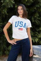 Woman wearing LIBERTY classic USA graphic crewneck baby tee featuring soft single jersey cotton stretch fabric, fitted short sleeve baby tee silhouette, clean cropped length, classic crew neckline, bold USA graphic across the front, American flag graphic detail below, and straight fitted hemline.