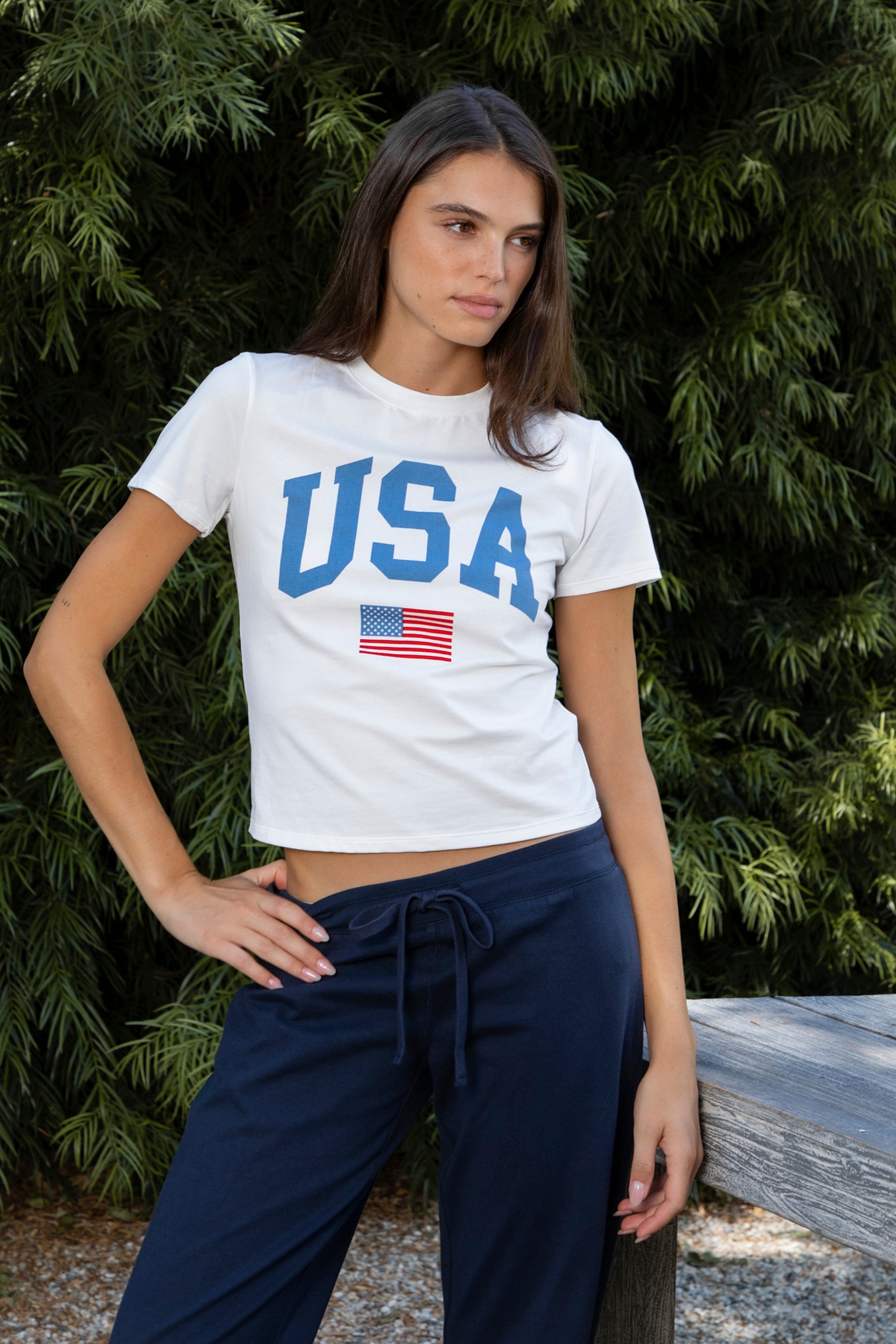 Woman wearing LIBERTY classic USA graphic crewneck baby tee featuring soft single jersey cotton stretch fabric, fitted short sleeve baby tee silhouette, clean cropped length, classic crew neckline, bold USA graphic across the front, American flag graphic detail below, and straight fitted hemline.