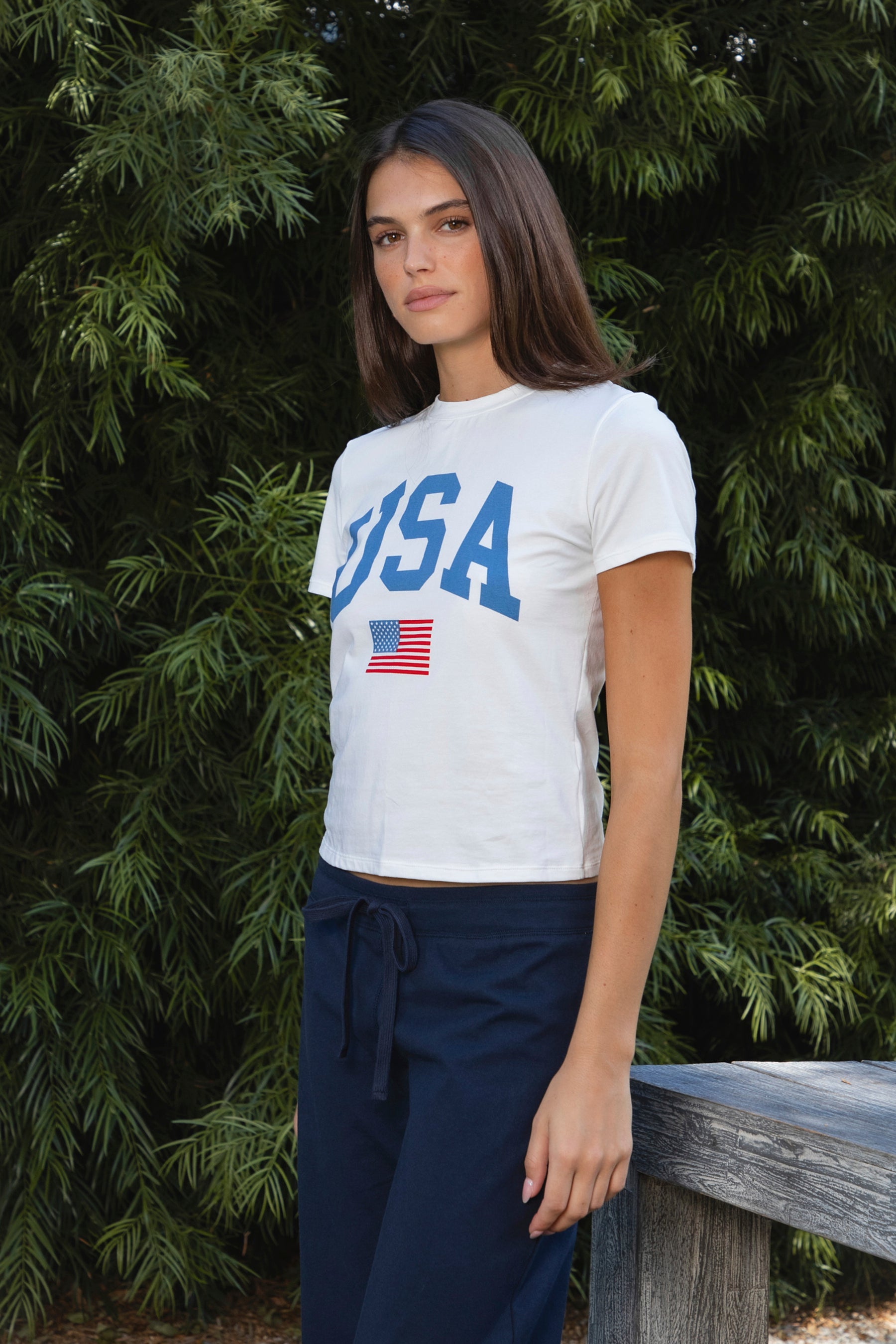 Woman wearing LIBERTY classic USA graphic crewneck baby tee featuring soft single jersey cotton stretch fabric, fitted short sleeve baby tee silhouette, clean cropped length, classic crew neckline, bold USA graphic across the front, American flag graphic detail below, and straight fitted hemline.