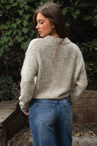 Woman wearing TIC TOC TF31840 SELYNA heather oatmeal knit pullover sweater with a soft wool-blend texture, relaxed oversized fit, drop shoulders, and long ribbed-cuff sleeves paired with medium-wash denim jeans in a cozy outdoor garden setting.