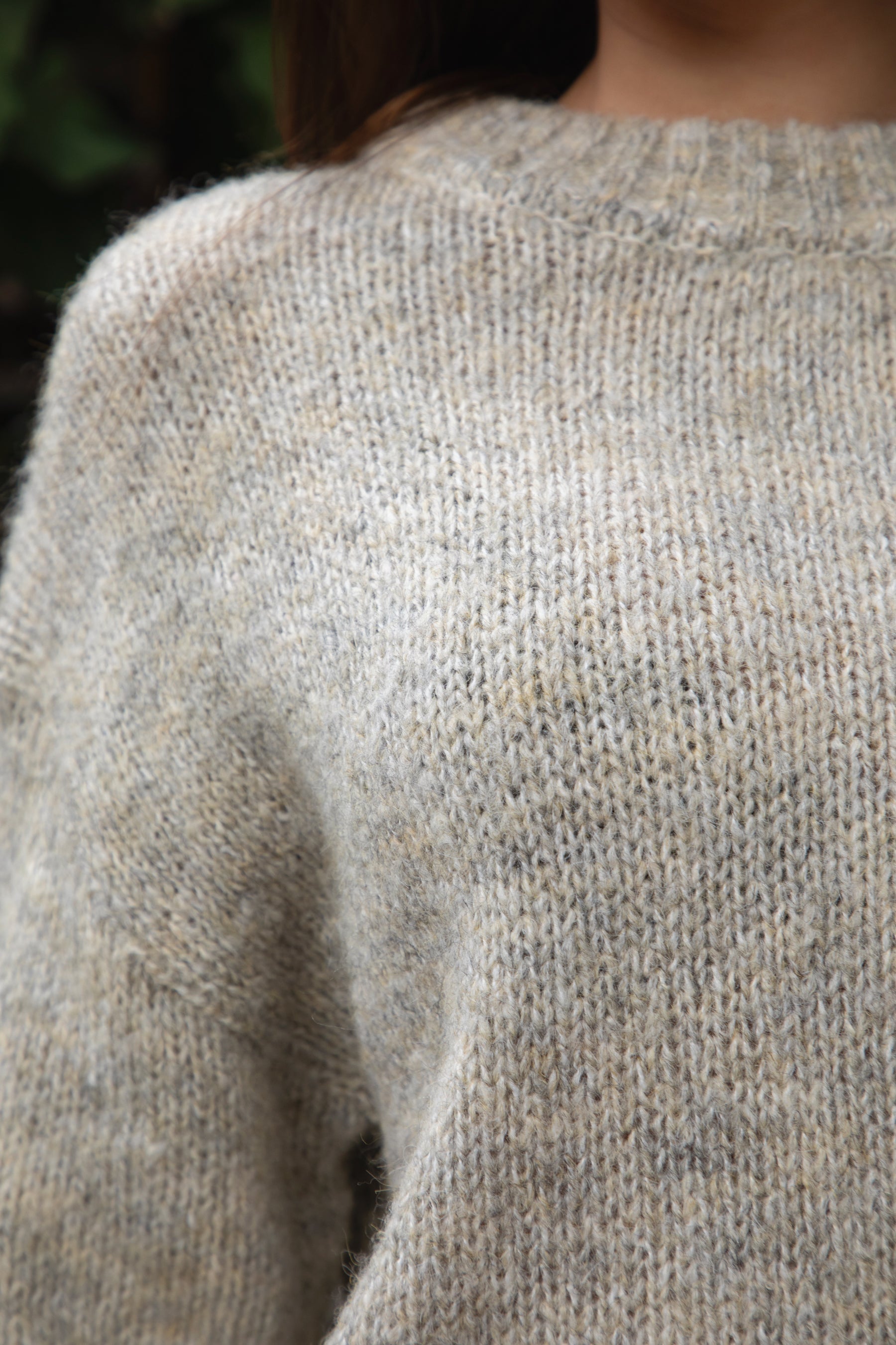 Woman wearing TIC TOC TF31840 SELYNA heather oatmeal knit pullover sweater with a soft wool-blend texture, relaxed oversized fit, drop shoulders, and long ribbed-cuff sleeves paired with medium-wash denim jeans in a cozy outdoor garden setting.