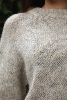 Woman wearing TIC TOC TF31840 SELYNA heather oatmeal knit pullover sweater with a soft wool-blend texture, relaxed oversized fit, drop shoulders, and long ribbed-cuff sleeves paired with medium-wash denim jeans in a cozy outdoor garden setting.