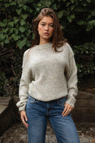 Woman wearing TIC TOC TF31840 SELYNA heather oatmeal knit pullover sweater with a soft wool-blend texture, relaxed oversized fit, drop shoulders, and long ribbed-cuff sleeves paired with medium-wash denim jeans in a cozy outdoor garden setting.