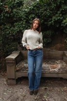 Woman wearing TIC TOC TF31840 SELYNA heather oatmeal knit pullover sweater with a soft wool-blend texture, relaxed oversized fit, drop shoulders, and long ribbed-cuff sleeves paired with medium-wash denim jeans in a cozy outdoor garden setting.