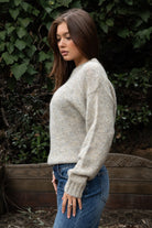 Woman wearing TIC TOC TF31840 SELYNA heather oatmeal knit pullover sweater with a soft wool-blend texture, relaxed oversized fit, drop shoulders, and long ribbed-cuff sleeves paired with medium-wash denim jeans in a cozy outdoor garden setting.