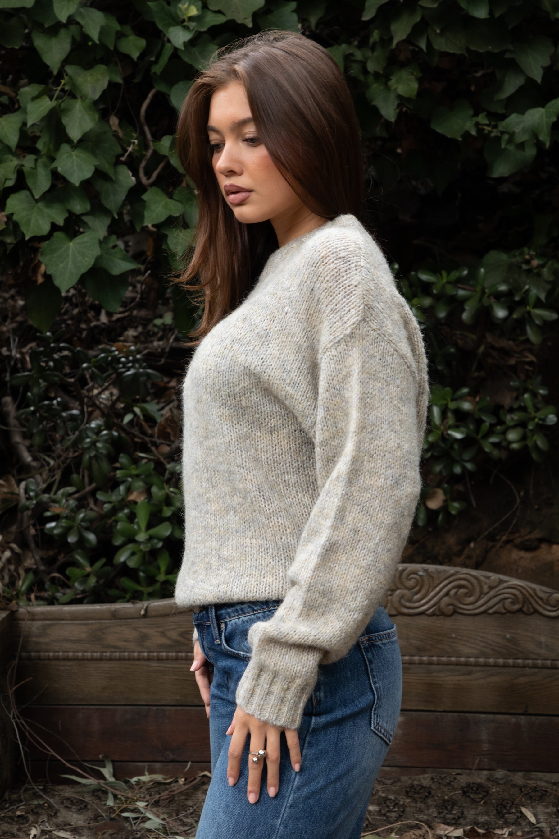 Woman wearing TIC TOC TF31840 SELYNA heather oatmeal knit pullover sweater with a soft wool-blend texture, relaxed oversized fit, drop shoulders, and long ribbed-cuff sleeves paired with medium-wash denim jeans in a cozy outdoor garden setting.