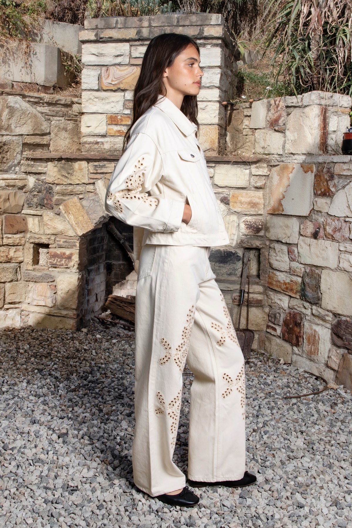 Woman wearing ALBA embroidered wide leg denim pants featuring structured cotton blend denim, high rise waist, classic four pocket design, intricate floral eyelet embroidery throughout the legs, relaxed wide leg silhouette, full length fit, button and zip fly closure, and belt loops along the waistline.