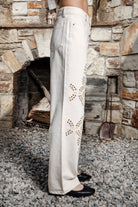 Woman wearing ALBA embroidered wide leg denim pants featuring structured cotton blend denim, high rise waist, classic four pocket design, intricate floral eyelet embroidery throughout the legs, relaxed wide leg silhouette, full length fit, button and zip fly closure, and belt loops along the waistline.