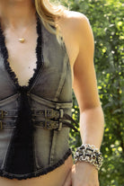 Woman wearing TIC TOC TG31902 CALEXA distressed washed denim halter top in a washed black denim finish, featuring a deep halter neckline, distressed frayed trim, front faux eyelet belt detail, contrast white stitching, cropped fitted silhouette, and low back zipper closure for an edgy western festival look.