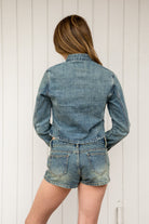 Woman wearing REMINGTON military semi crop denim jacket featuring cotton blend denim, structured tailored fit, military inspired horizontal strap panels, matte metal button accents, stand collar, hidden hook and eye front closure, long sleeves with metal button cuffs, and cropped hemline.
