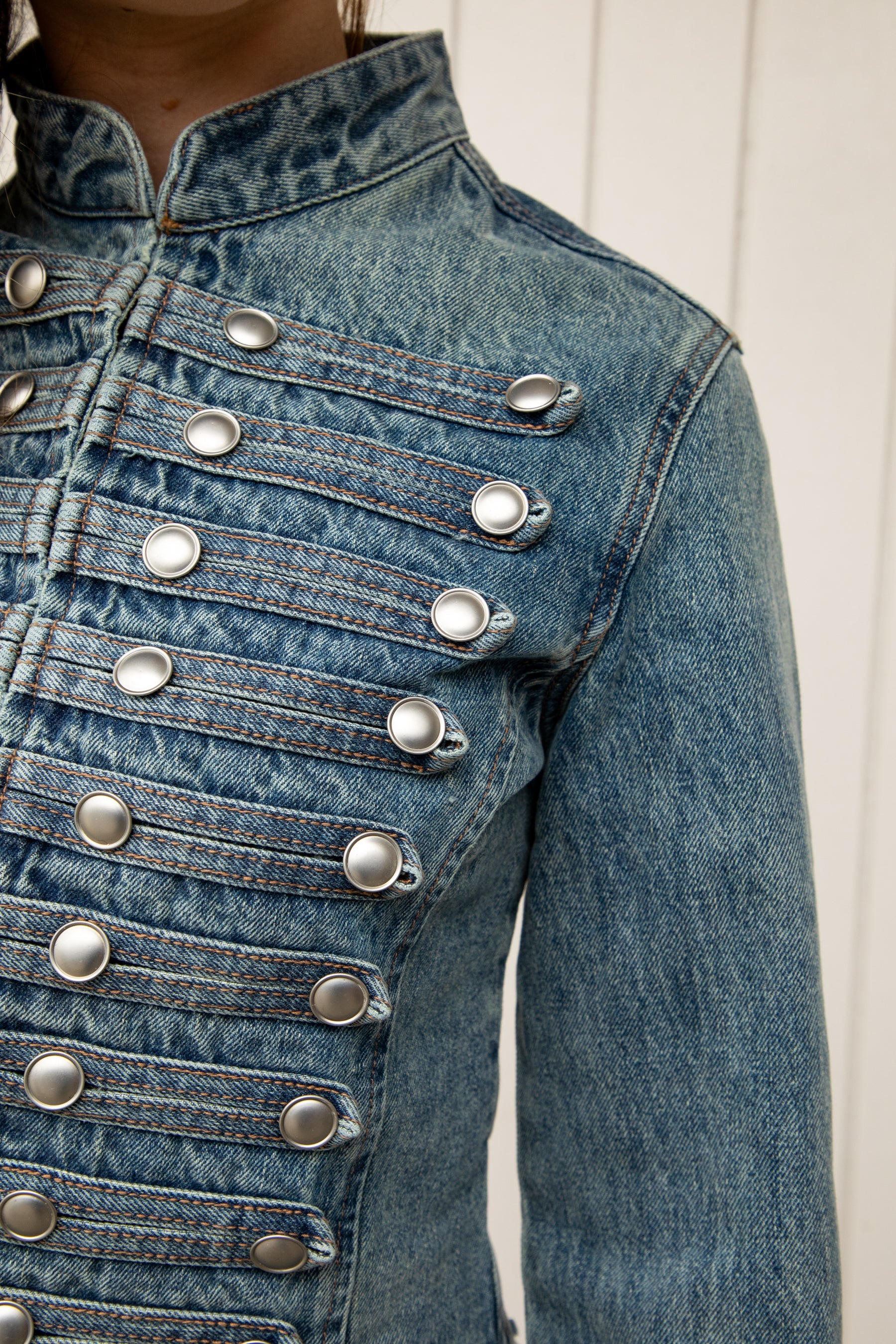 Woman wearing REMINGTON military semi crop denim jacket featuring cotton blend denim, structured tailored fit, military inspired horizontal strap panels, matte metal button accents, stand collar, hidden hook and eye front closure, long sleeves with metal button cuffs, and cropped hemline.