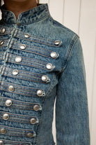 Woman wearing REMINGTON military semi crop denim jacket featuring cotton blend denim, structured tailored fit, military inspired horizontal strap panels, matte metal button accents, stand collar, hidden hook and eye front closure, long sleeves with metal button cuffs, and cropped hemline.