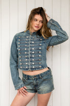 Woman wearing REMINGTON military semi crop denim jacket featuring cotton blend denim, structured tailored fit, military inspired horizontal strap panels, matte metal button accents, stand collar, hidden hook and eye front closure, long sleeves with metal button cuffs, and cropped hemline.