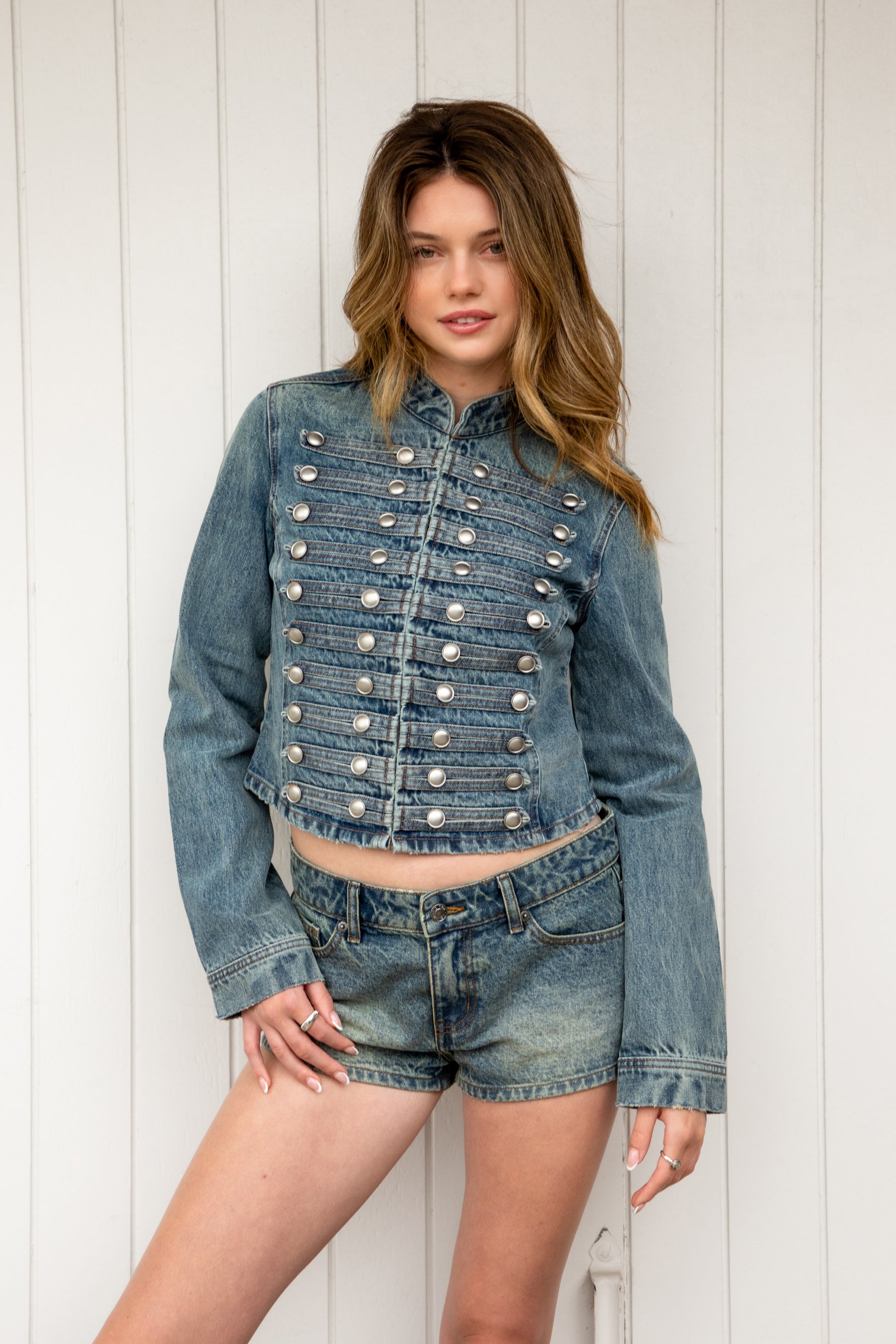 Woman wearing REMINGTON military semi crop denim jacket featuring cotton blend denim, structured tailored fit, military inspired horizontal strap panels, matte metal button accents, stand collar, hidden hook and eye front closure, long sleeves with metal button cuffs, and cropped hemline.