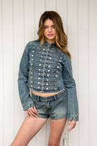 Woman wearing REMINGTON military semi crop denim jacket featuring cotton blend denim, structured tailored fit, military inspired horizontal strap panels, matte metal button accents, stand collar, hidden hook and eye front closure, long sleeves with metal button cuffs, and cropped hemline.