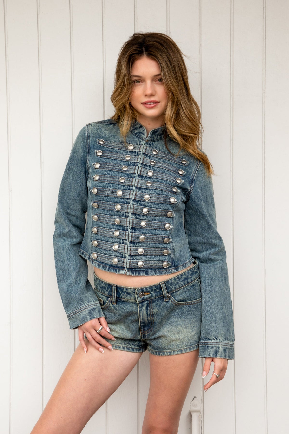 Woman wearing REMINGTON military semi crop denim jacket featuring cotton blend denim, structured tailored fit, military inspired horizontal strap panels, matte metal button accents, stand collar, hidden hook and eye front closure, long sleeves with metal button cuffs, and cropped hemline.