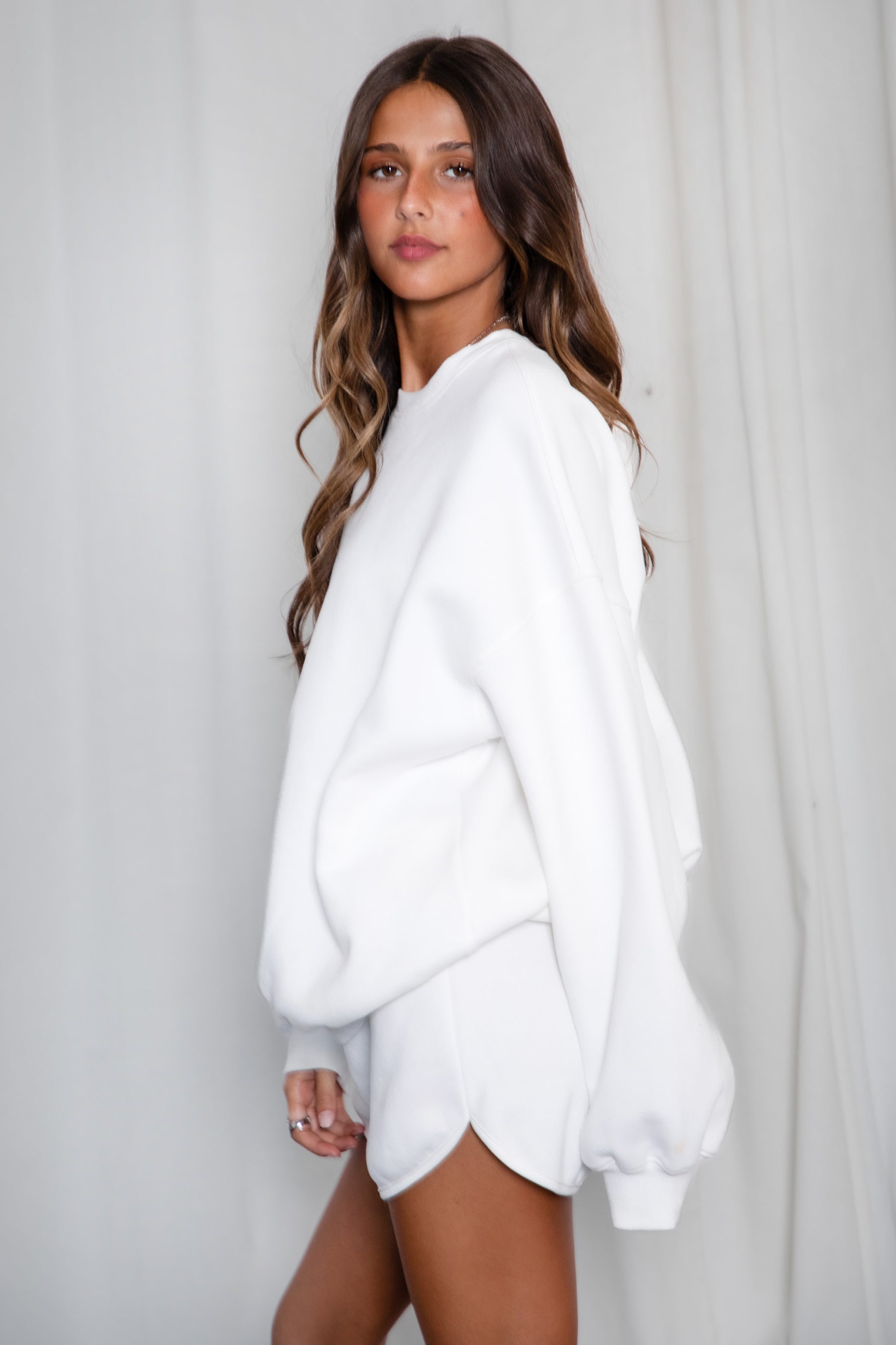 Woman wearing a white oversized sweater and shorts against a plain background