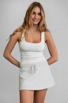 Woman wearing TIC TOC SOMI Core Basic Rib Jersey Tank Top in heather white with low square neckline, wide straps, and form-fitting ribbed fabric, paired with a heather white mini skirt.