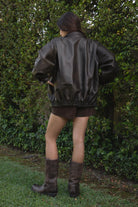 Women wearing TIC TOC GIANA Oversized PU Leather Bomber Jacket in dark brown with collared neckline, puff sleeves, and elastic waistband over a white baby doll floral top, suede lace up shorts, and brown frye boots standing against a green leafy background. 