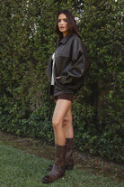 Women wearing TIC TOC GIANA Oversized PU Leather Bomber Jacket in dark brown with collared neckline, puff sleeves, and elastic waistband over a white baby doll floral top, suede lace up shorts, and brown frye boots standing against a green leafy background. 