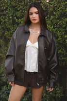 Women wearing TIC TOC GIANA Oversized PU Leather Bomber Jacket in dark brown with collared neckline, puff sleeves, and elastic waistband over a white baby doll floral top and suede lace up shorts, standing against a green leafy background. 